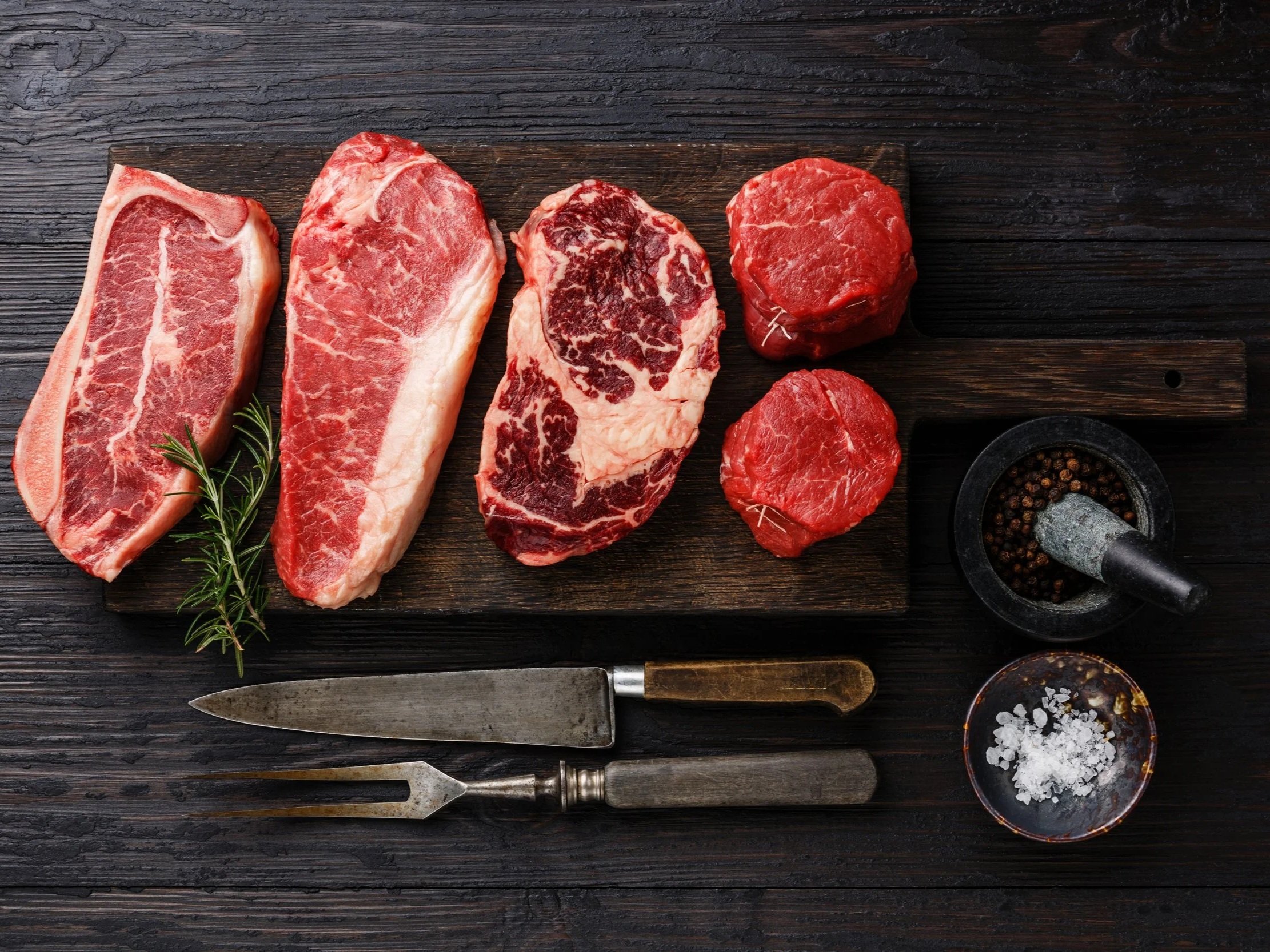 Fresh cuts of beef steaks including ribeye, sirloin, and tenderloin arranged on a wooden cutting board, accompanied by kitchen tools, a mortar and pestle with peppercorns, and a bowl of sea salt, garnished with a sprig of rosemary.