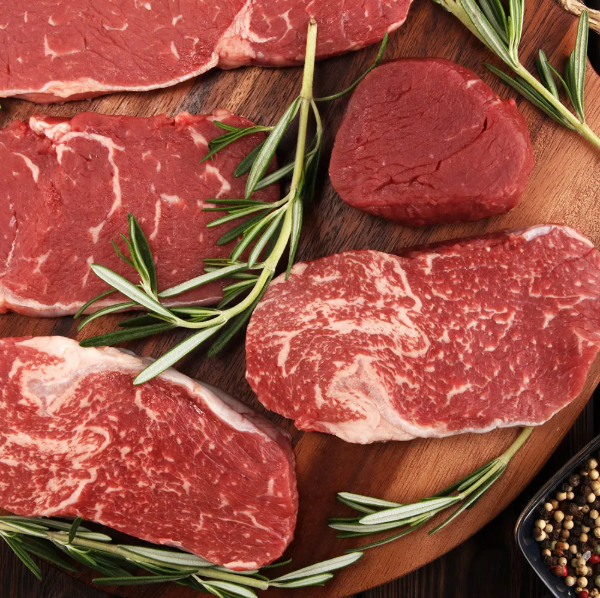 Fresh cuts of beef steak, including ribeye and tenderloin, arranged on a wooden cutting board with sprigs of rosemary and peppercorns, showcasing high-quality marbling and vibrant red color.