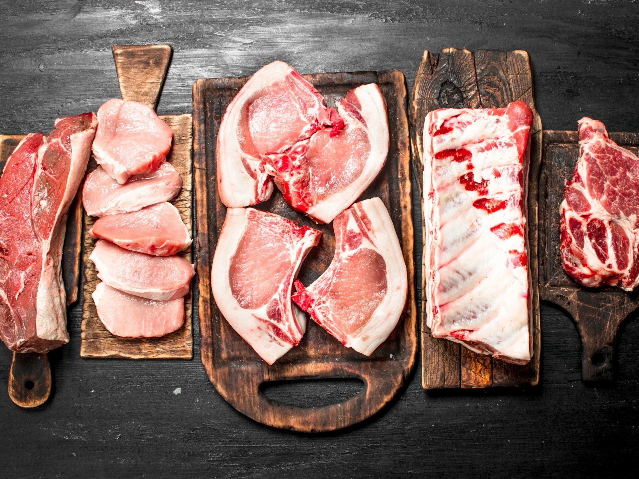 Assorted cuts of raw meat displayed on wooden cutting boards, including pork chops, pork loin, and ribs, set against a dark textured background. Ideal for culinary and butchery references.